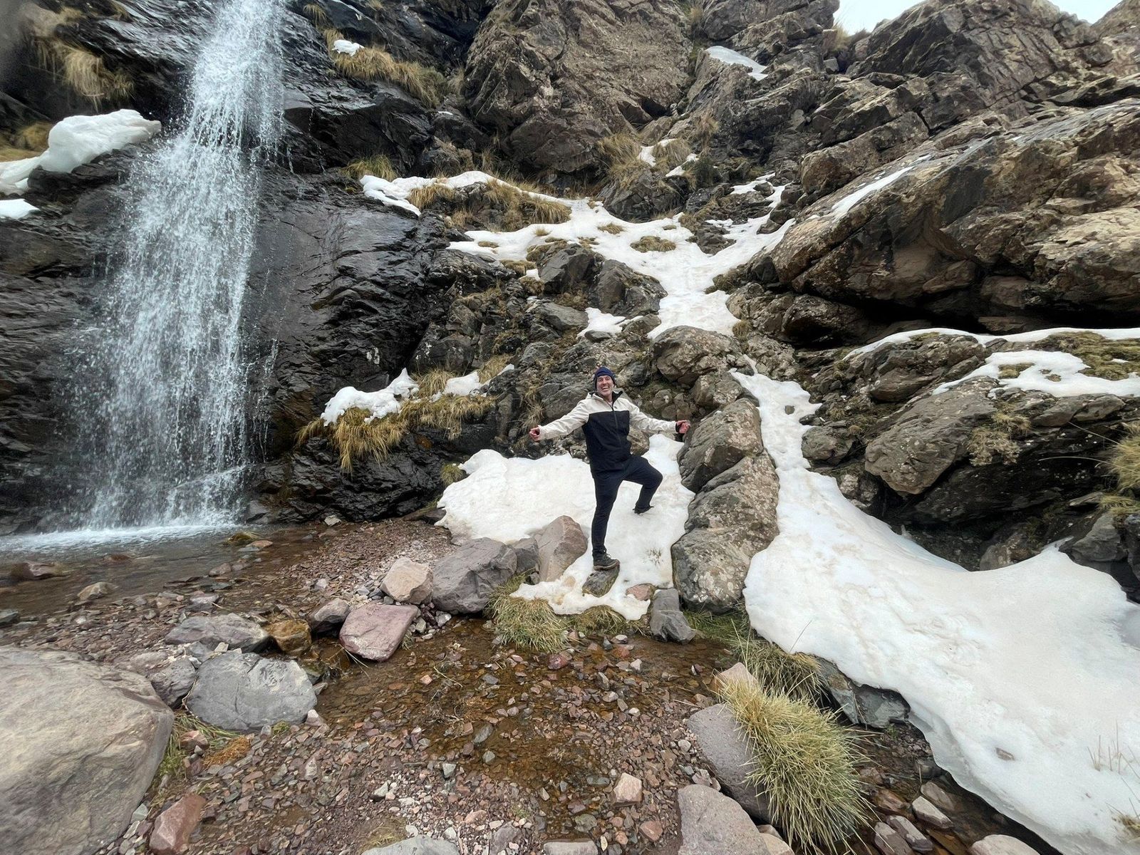 A hiker smiling next to a snow-bordered waterfall in High Atlas Mountains