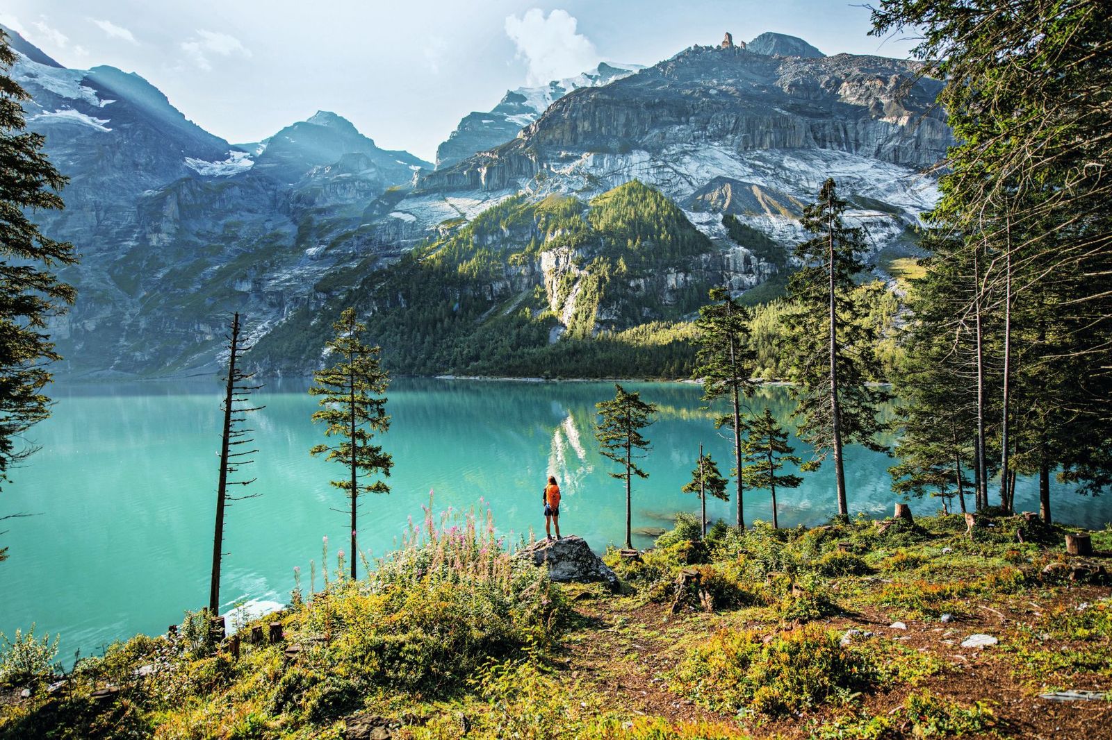 A hiker with a backpack stands on a rocky outcrop overlooking the turquoise waters of Lake Oeschinen