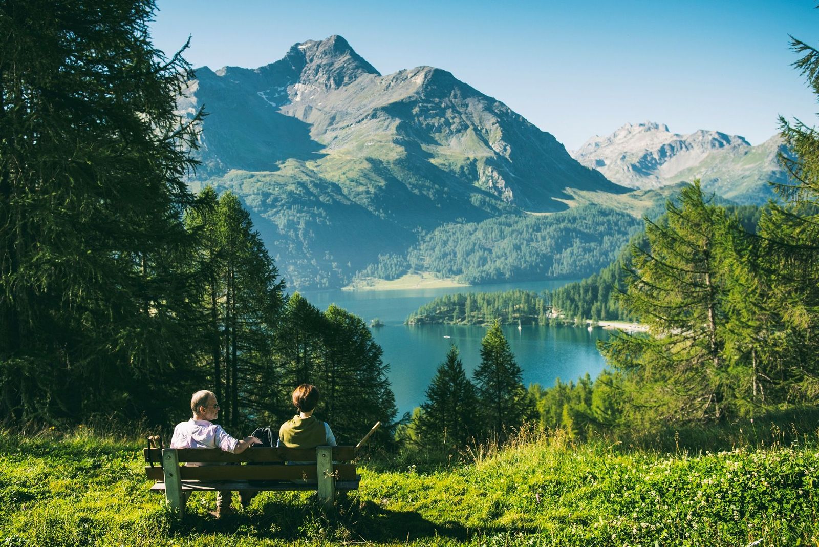 Two hikers resting on a wooden bench looking out over the clear blue waters of Lake Sils