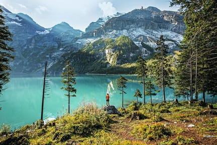 A hiker with a backpack stands on a rocky outcrop overlooking the turquoise waters of Lake Oeschinen